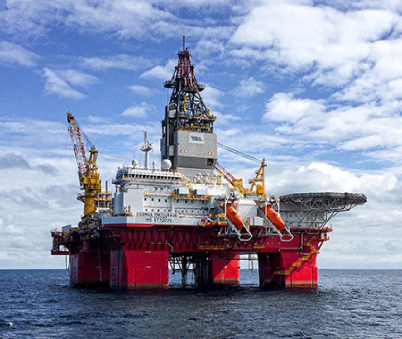 Large red offshore oil drilling platform with crane equipment in open ocean under blue sky with clouds